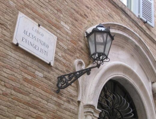 Targa stradale "Largo Alessandro Evangelista" affissa su un edificio in laterizio, con un lampione decorativo in ferro battuto accanto all'arco di un portone.