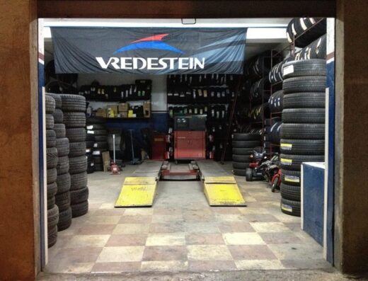 Interno di un'officina di riparazione moto con ponte elevatore e montagne di pneumatici. Banner Vredestein in alto. Accessori e ricambi per moto visibili.