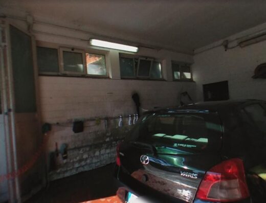 Interno di un garage con una Toyota Yaris verde scuro parcheggiata. L'ambiente è illuminato da una luce al neon e presenta finestre in alto. La porta è aperta.