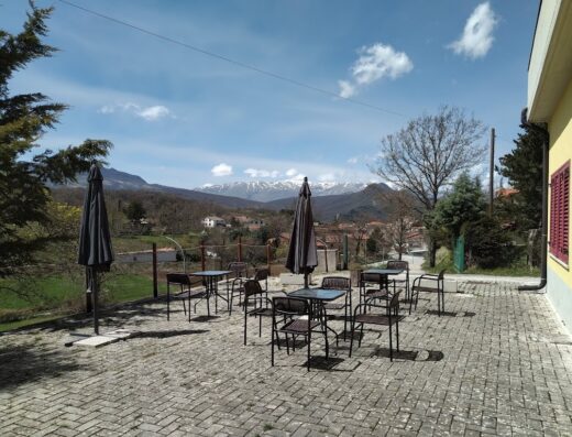 Esterno del Rifugio dei Sanniti con tavoli, sedie e ombrelloni. sullo sfondo montagne innevate e cielo azzurro, ideale per motociclisti in viaggio.