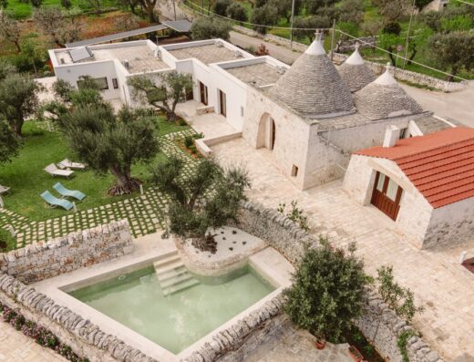 Visione aerea di un rifugio di Puglia, un complesso di trulli e dimore con piscina, giardino curato e architettura tradizionale in pietra.