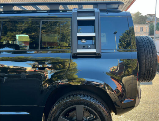 Dettaglio di una Land Rover Defender nera con accessori per il viaggio: ruota di scorta posteriore, scaletta per il tetto e barre portatutto, ideale per l'autolavaggio Risto-Rally a Genova Aeroporto.