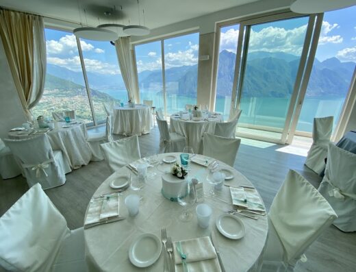 Ristorante Hotel Panoramico: Elegante sala da pranzo con tavoli imbanditi in bianco, vista mozzafiato su lago e montagne, ideale per matrimoni e eventi speciali.