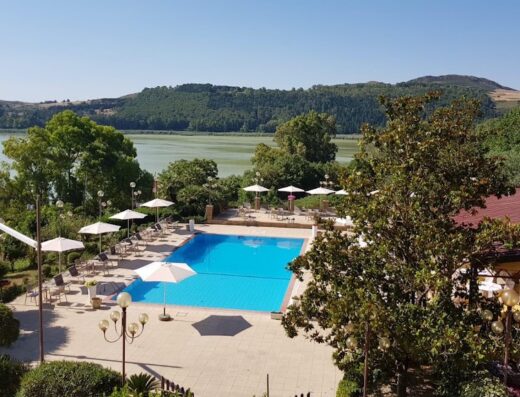 Panoramic view of the Riviera Hotel featuring a bright blue swimming pool, sun umbrellas, and a serene lake backdrop.