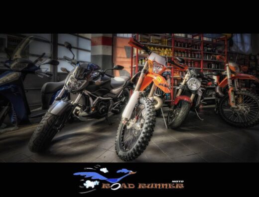 Esposizione di diverse moto, tra cui una moto da cross arancione, una moto da strada Honda e uno scooter blu, all'interno di un'officina Road Runner Moto con scaffali di ricambi e accessori.