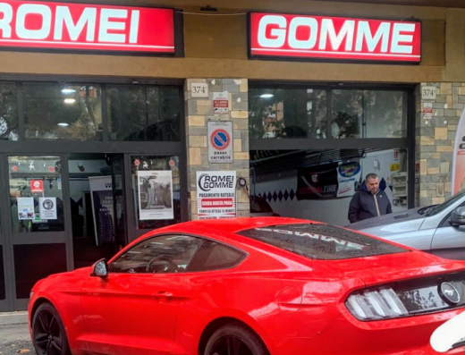 Foto del negozio Romei Gomme con auto rossa parcheggiata di fronte. Servizi professionali per pneumatici auto e moto, accessori e vendita. Manutenzione e riparazione gomme.