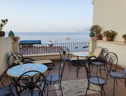 Balcone panoramico a Romeo e Giulietta con tavoli decorati, sedie in ferro battuto e vista sul mare con barche.