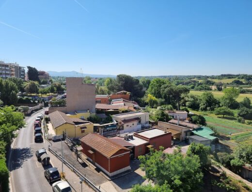 Vista panoramica dell'Hotel Romoli, con strada alberata, case colorate e campagna sullo sfondo, in una giornata di sole.