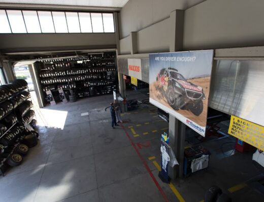 Interno dell'officina Rossi Gomme con scaffali di pneumatici, cartello Maxxis e banner "Are you driver enough?" con auto da rally BFGoodrich, servizio sostituzione gomme moto e auto.