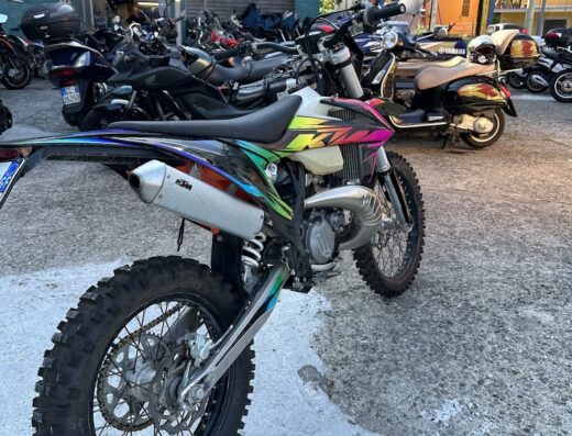 Una moto KTM da cross con dettagli colorati parcheggiata di fronte a un negozio di moto con altre moto e scooter visibili. Ideale per viaggi avventurosi e servizi di riparazione.