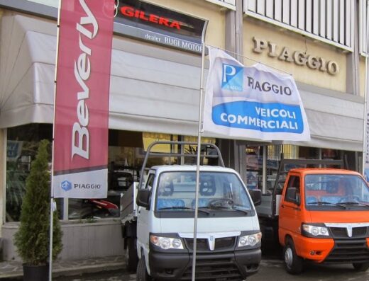 Rugi Motori dealership exterior featuring Piaggio commercial vehicles, including a white and an orange truck, and flags for Beverly and Piaggio Veicoli Commerciali.