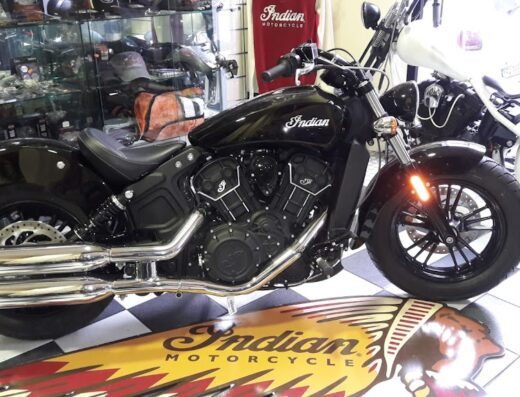 Una moto Indian nera in esposizione su un tappeto con il logo Indian Motorcycle. Accessori moto e altre moto visibili sullo sfondo.