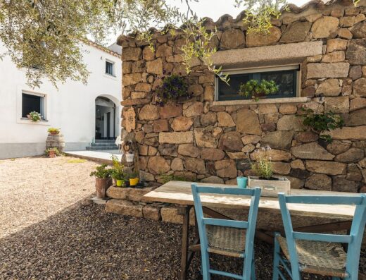 Esterno di Sa Crai B&B in Sardegna con tavolo e sedie in legno, muro in pietra e casa bianca sullo sfondo. Esperienza sarda.