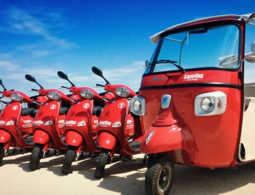 Line of red Sametour scooters and an Ape Calessino parked outdoors under a blue sky, ideal for motorcycle tours and rentals.