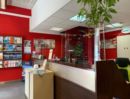 Interno di agenzia viaggi Sardaviaggi con bancone reception protetto da pannello in plexiglass, brochure turistiche esposte e logo Alitalia