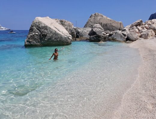 Una donna fa il bagno nelle acque cristalline di una spiaggia sarda, con rocce bianche e yacht sullo sfondo. Ideale per vacanze in barca.