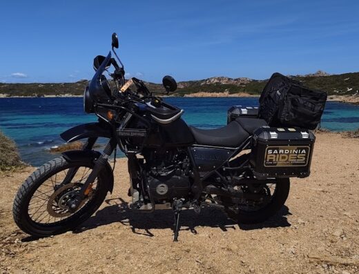 Motocicletta Royal Enfield accessoriata per il mototurismo in Sardegna, con bauletti e borsa, sullo sfondo il mare cristallino e le rocce dell'isola, offerta di Sardinia Rides.