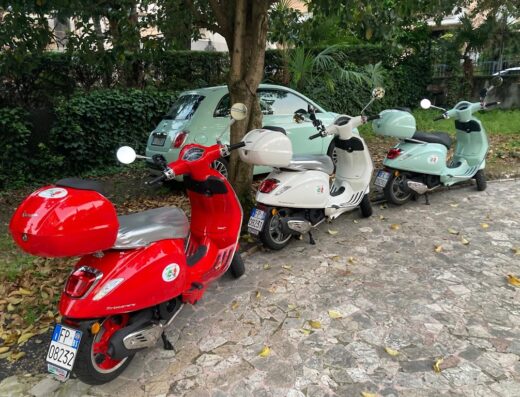 Noleggio scooter Vespa e auto a Desenzano: fila di Vespa Primavera rosse, bianche e turchesi parcheggiate su pavimentazione esterna.