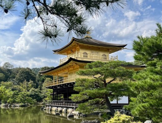 A golden pavilion reflected in a serene pond, framed by lush greenery and a partly cloudy sky, capturing the essence of Japanese travel and cultural heritage.