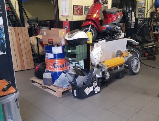 Un caotico angolo di un'officina motociclistica mostra uno scooter rosso appoggiato su un'altra moto, circondato da accessori, ricambi, bidoni di olio e attrezzi.