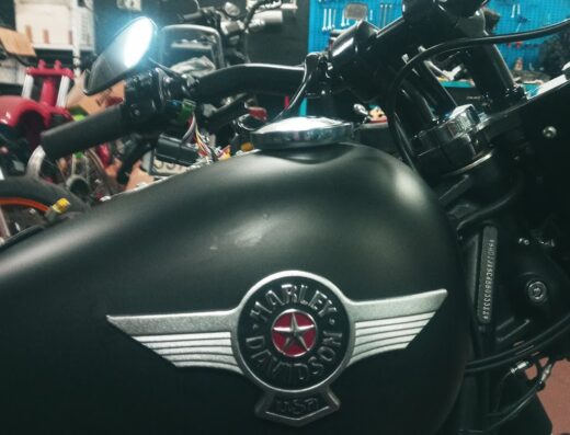 Serbatoio nero opaco di una motocicletta Harley-Davidson in un'officina, con logo iconico ben visibile. Dettagli del manubrio, accessori e altri veicoli sullo sfondo.