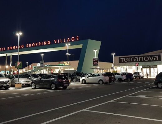 Parcheggio illuminato del Castel Romano Shopping Village di notte, con auto parcheggiate e insegne dei negozi visibili.