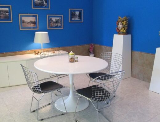 Interno di un locale con tavolo rotondo bianco circondato da sedie in metallo, credenza bianca, lampada, vasi decorativi e ceramiche artistiche raffiguranti teste siciliane su piedistalli bianchi, quadri appesi alla parete blu.