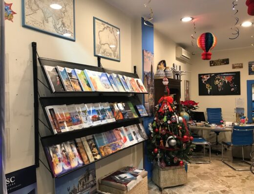 Interno dell'agenzia viaggi Siciliamando Viaggi di Viaggi di Gusto s.c.a.r.l. con scaffale di cataloghi turistici, albero di Natale decorato e mappe geografiche appese al muro.