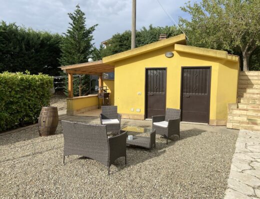 Area relax esterna in Sicilia con mobili da giardino in vimini, tavolo basso e sfondo di casetta gialla con due porte.