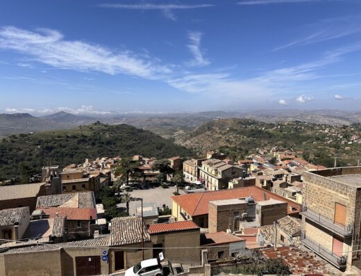 Veduta panoramica di un centro storico in Sicilia, con case dai tetti rossi e colline sullo sfondo, offrendo scorci tipici per un soggiorno autentico.