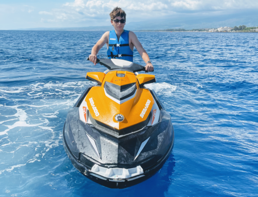 Ragazzo che guida una moto d'acqua Sea-Doo gialla e nera in mare aperto, con giubbotto di salvataggio e occhiali da sole, sullo sfondo costa siciliana.