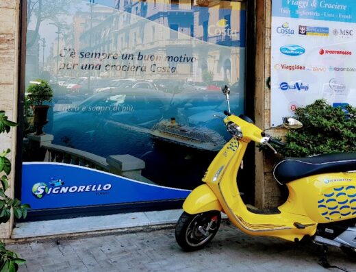 Signorello Viaggi: Vespa gialla parcheggiata di fronte all'agenzia con vetrina che pubblicizza crociere Costa, ideale per pianificare viaggi e vacanze.