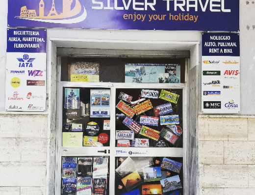 Immagine dell'agenzia viaggi Silver Travel, con insegna che mostra monumenti come il Colosseo e la Torre Eiffel. La vetrina è piena di adesivi e cartoline di varie destinazioni turistiche, con offerte di biglietteria aerea, marittima e ferroviaria, e servizi di noleggio auto e moto.
