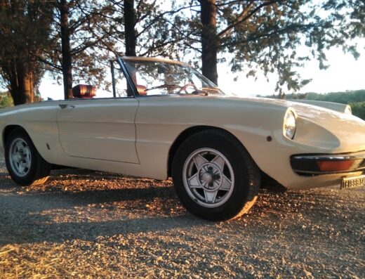 Alfa Romeo Spider d'epoca color crema su strada sterrata, ideale per un viaggio slow in Italia, tra accessori e servizi esclusivi.