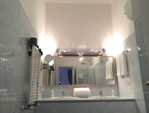 Bagno luminoso con piastrelle azzurre, doppio lavabo, specchio ampio, asciugamani bianchi e scaldasalviette a parete presso SmooRoomS Ostiense.