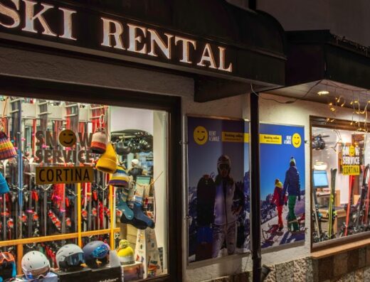Vetrine di un negozio di noleggio sci a Cortina, con sci, scarponi, caschi e accessori esposti, illuminato da luci festive. Servizi invernali.