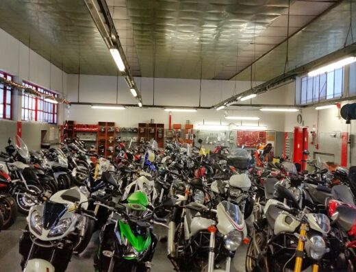 Interno di un negozio o officina con molte moto parcheggiate. Moto di diversi marchi e modelli, accessori e attrezzature visibili. Servizio di vendita e riparazione moto.