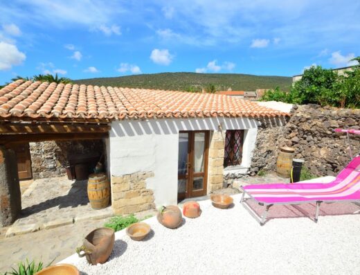 Esterno di S'Ommu Ezza, edificio rurale con tetto in tegole, pareti bianche e dettagli in pietra. Presente una zona relax con lettino rosa e vasi di terracotta decorativi.