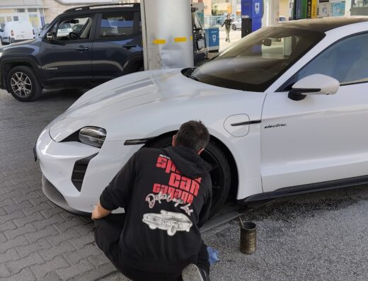 Un uomo accovacciato pulisce una Porsche Taycan bianca in una stazione di servizio. L'uomo indossa una felpa con cappuccio 'Speed Car Garage'.