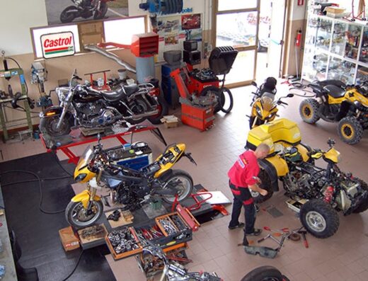 Officina SpeedMax affollata con diverse moto e quad in riparazione, meccanico al lavoro, accessori e ricambi visibili, insegna Castrol.