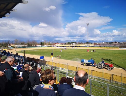 Immagine dello Speedway Moto Club Olimpia Terenzano: pista di speedway con trattore che innaffia la pista, tribune piene di spettatori e sullo sfondo le montagne