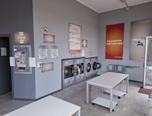Interno di Speedy Wash Lavanderia Self Service con lavatrici a gettoni, cassa automatica, tavoli pieghevoli e sedie per clienti.