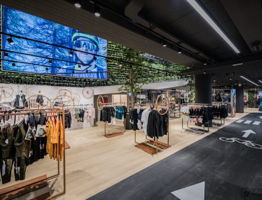 Interior of SPORTLER bike store featuring cycling apparel displayed on racks, a large screen showing a cyclist, and bike lane markings on the floor.