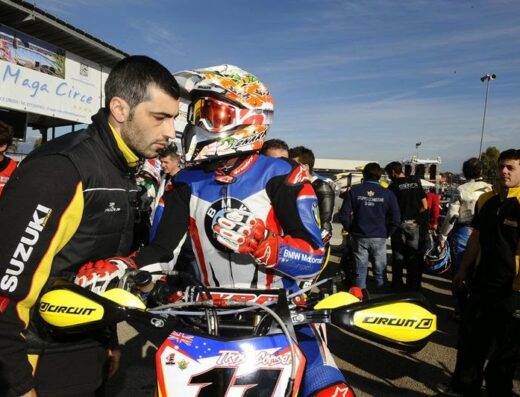 Immagine di ST Racing che mostra un pilota motociclistico con casco Shark e tuta BMW Motosport su una moto con protezioni Circuit gialle, assistito da un meccanico Suzuki in un evento di corsa.