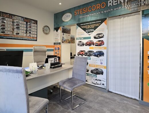 Interni di Stesicoro Rent, agenzia di noleggio auto, scooter e moto. In primo piano, bancone con computer e sedie. Sullo sfondo, cartelli con listino prezzi e informazioni sui servizi di autonoleggio e motonoleggio.