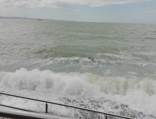 Onde che si infrangono sulla costa a Strada del Friuli 453, Contovello, con vista sul mare e orizzonte nuvoloso. Paesaggio costiero con ringhiera.