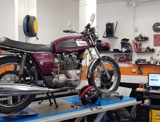 A maroon Triumph Trident motorcycle on a lift at StudioMotoRoma, a motorcycle service and repair shop, with mechanic in background.