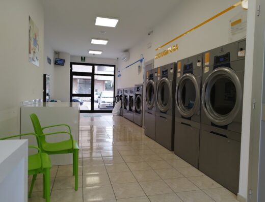 Interno di una lavanderia self-service Sun Wash con lavatrici industriali grigie allineate, pavimenti piastrellati e sedie verdi per l'attesa.