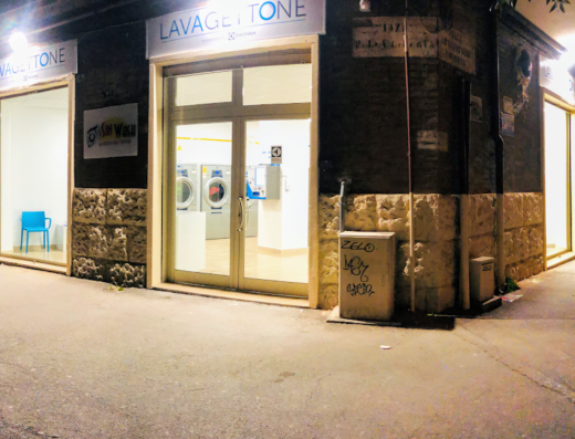 Sun Wash s.r.l. Lavagettone at night: a laundromat with washing machines visible through the glass door and a blue chair in the waiting area, offering self-service laundry.