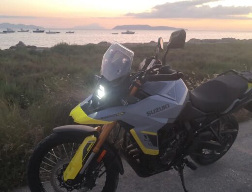Una moto Suzuki V-Strom 800DE grigia e gialla parcheggiata vicino alla riva, con vista sul mare al tramonto e barche ormeggiate.